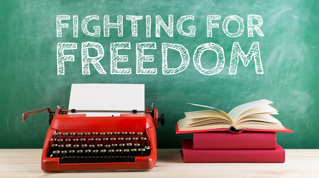 A typewriter and a stack of books in front of a chalkboard with words that read "FIGHTING FOR FREEDOM"