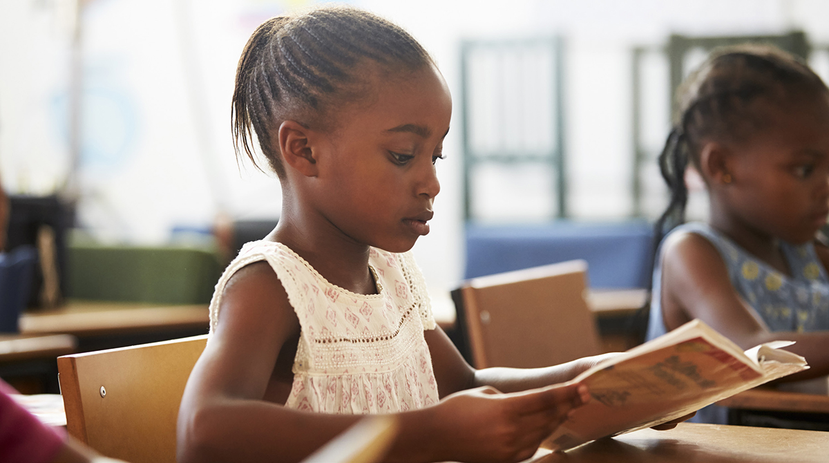 A young Black girl reads from a book.