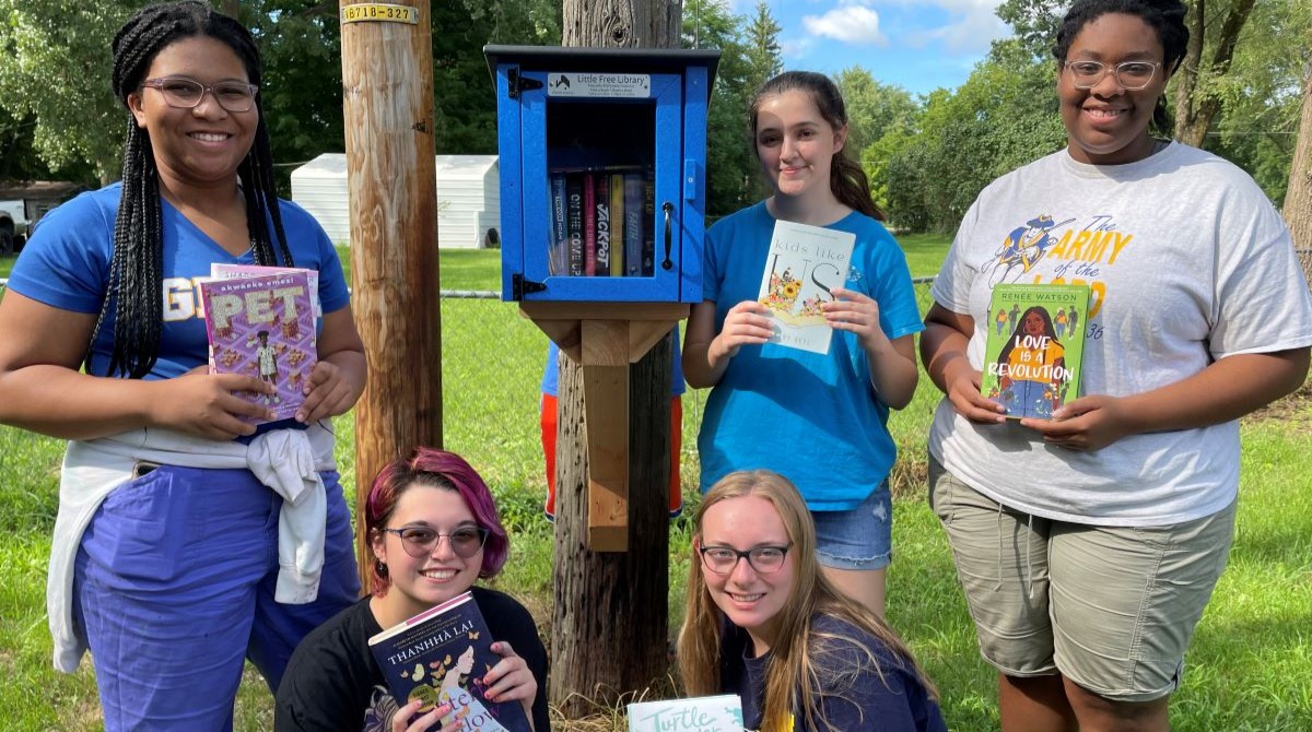 Photo of five young people holding books and posing with a Little Free Library.