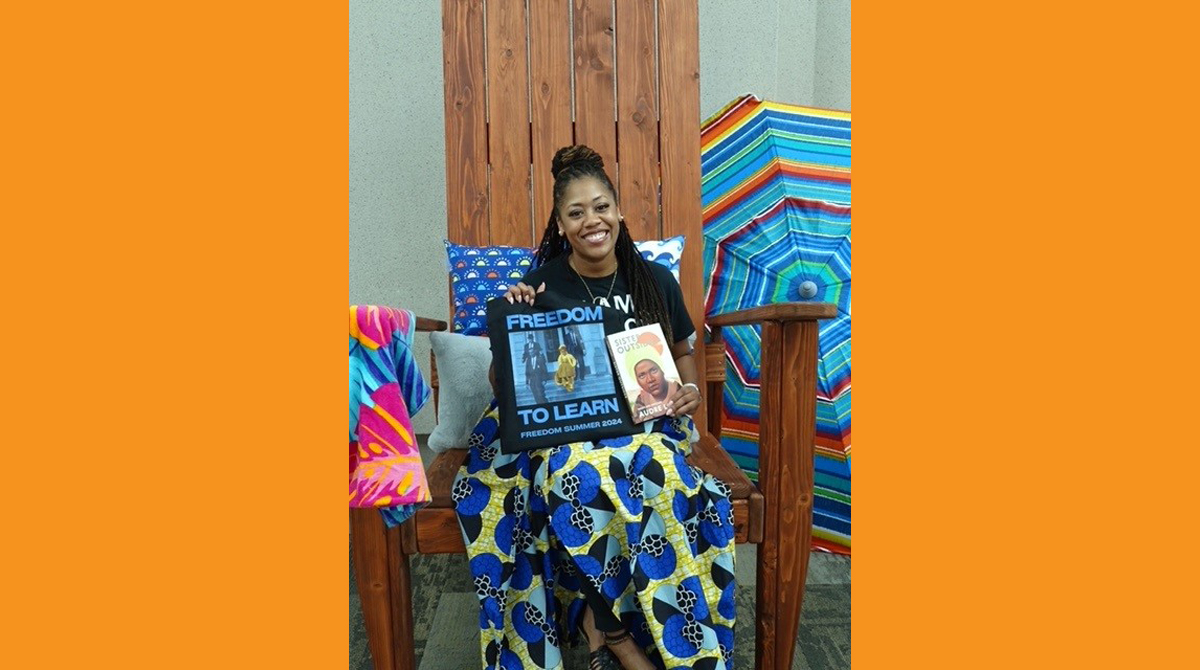 Photo of a woman - Dr. Ashley Gibson - shown seated in the Big Chair, holding a copy of "Sister Outsider: Essays and Speeches" by Audre Lorde and a FREEDOM TO LEARN tote bag.