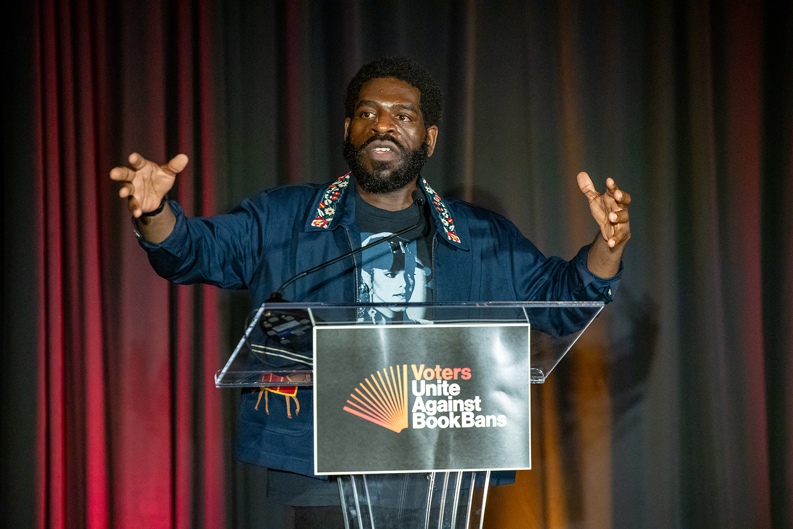 Hanif Abdurraqib speaking at a podium with a sign that says "VOTERS UNITE AGAINST BOOK BANS"