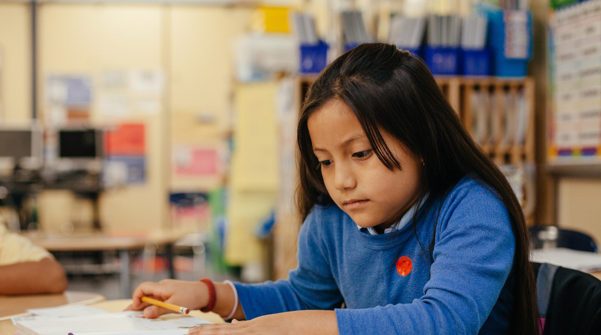 A child sits in a classroom looking at a book and holding a pencil.