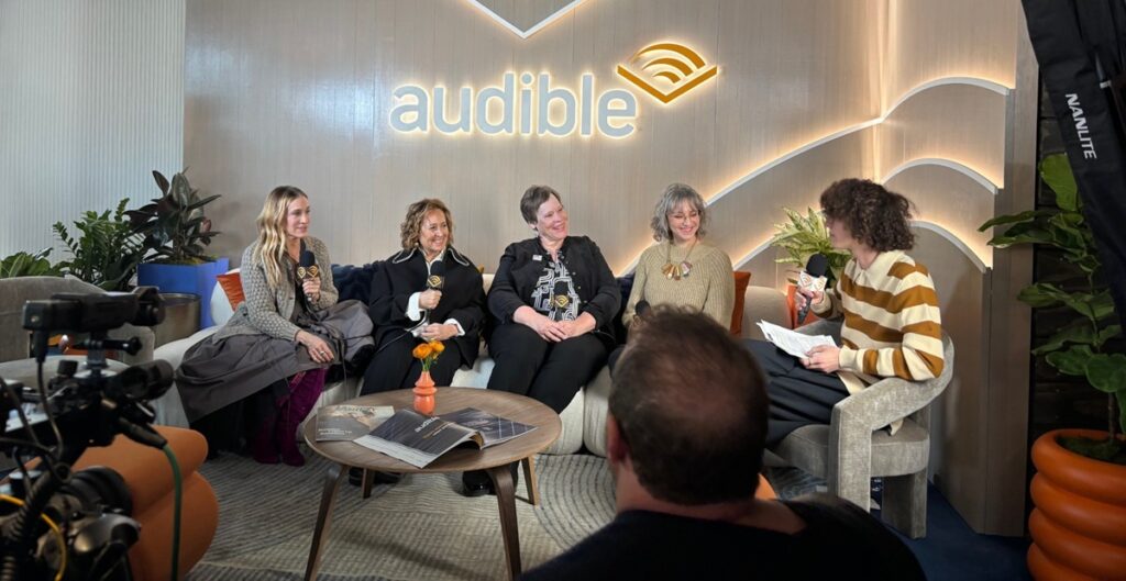 (From left to right) Sarah Jessica Parker, Kim Snyder, Martha Hickson, Audrey Wilson-Youngblood, and an interviewer sit together for Audible podcast recording about The Librarians. Audible's logo appears in the background and a camera is shown at the left.