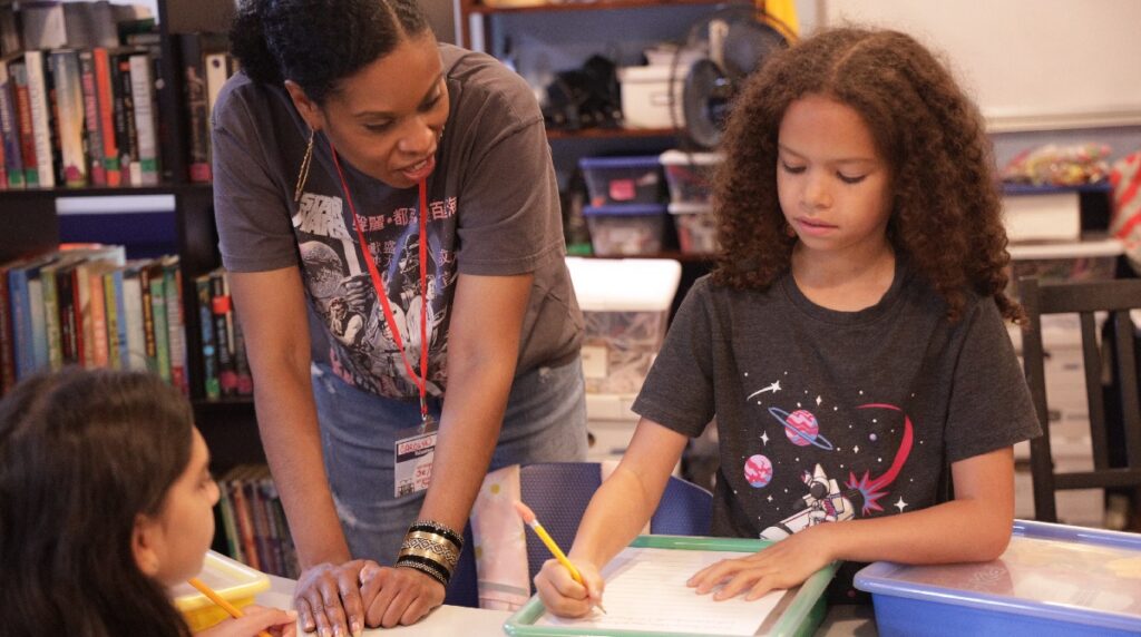 Two students are shown in a classroom or library setting. They are holding pencils and paper, and a teacher is leaning down in conversation with them.